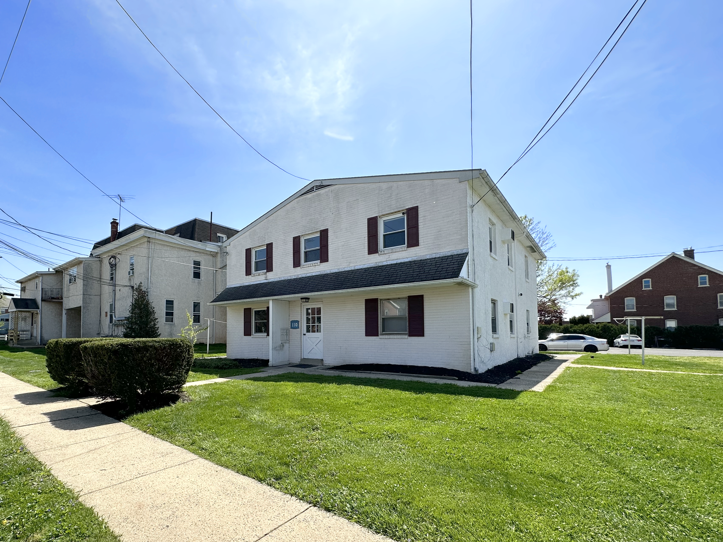 Two-story white building with red shutters, surrounded by green grass and walkways, in a residential neighborhood under a clear blue sky.