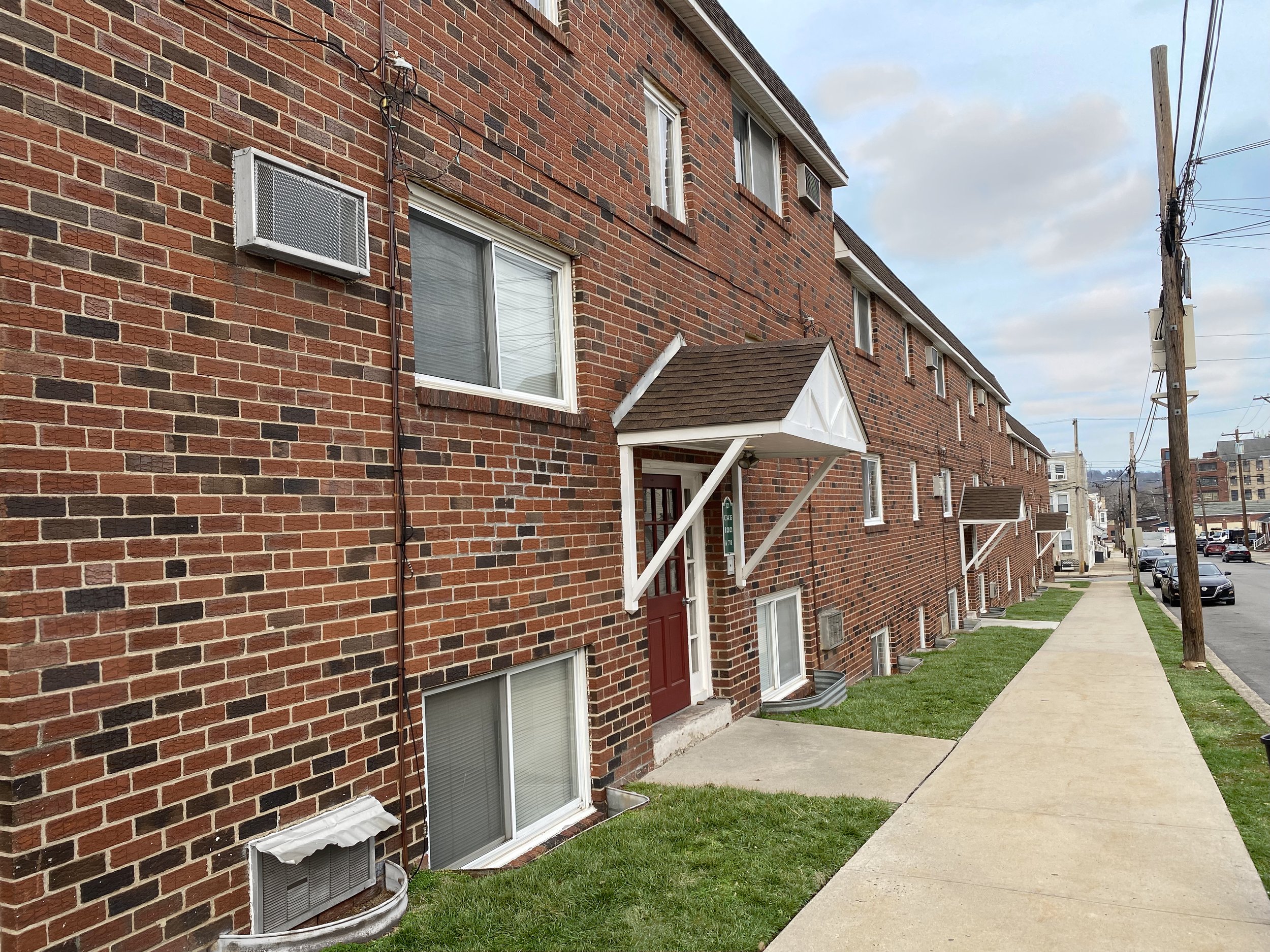 Brick-fronted apartment building with awning-covered entrances along a sidewalk, showcasing air conditioning units and greenery.