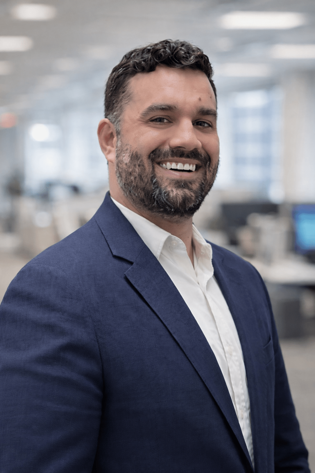 Smiling man in professional attire stands in an office setting, conveying confidence and approachability. The background features blurred office elements.