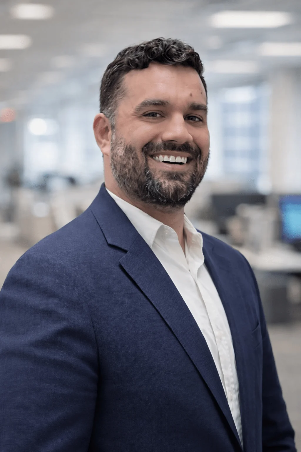 Smiling man in professional attire stands in an office setting, conveying confidence and approachability. The background features blurred office elements.