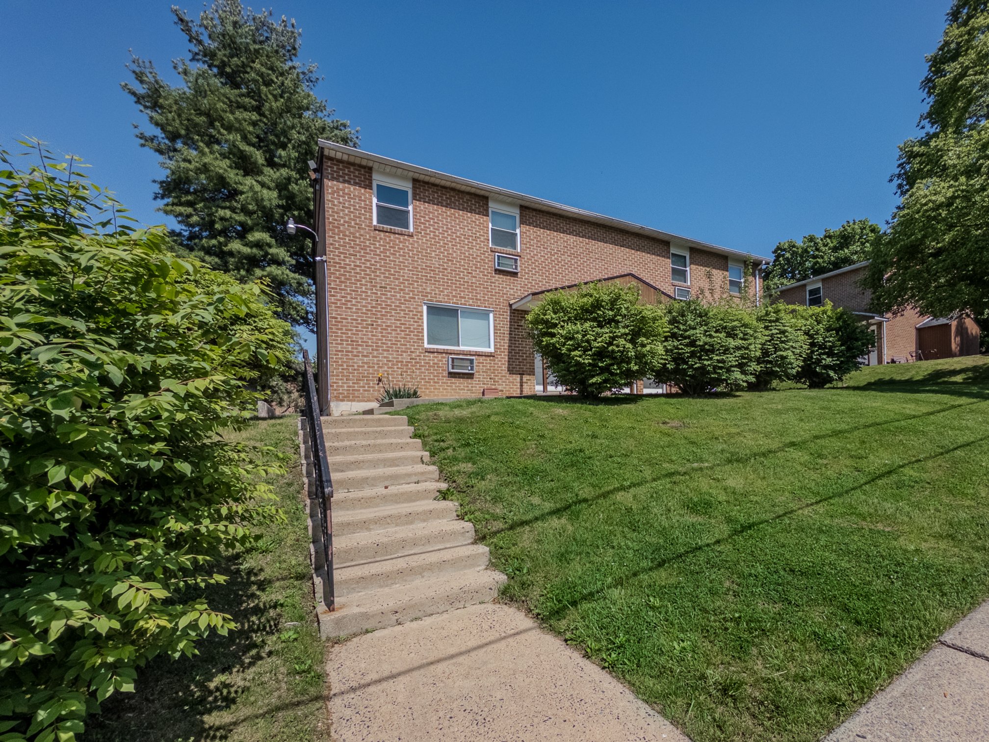 Two-story brick apartment building with stairs leading up to the entrance, surrounded by green lawns and shrubs under a clear blue sky.