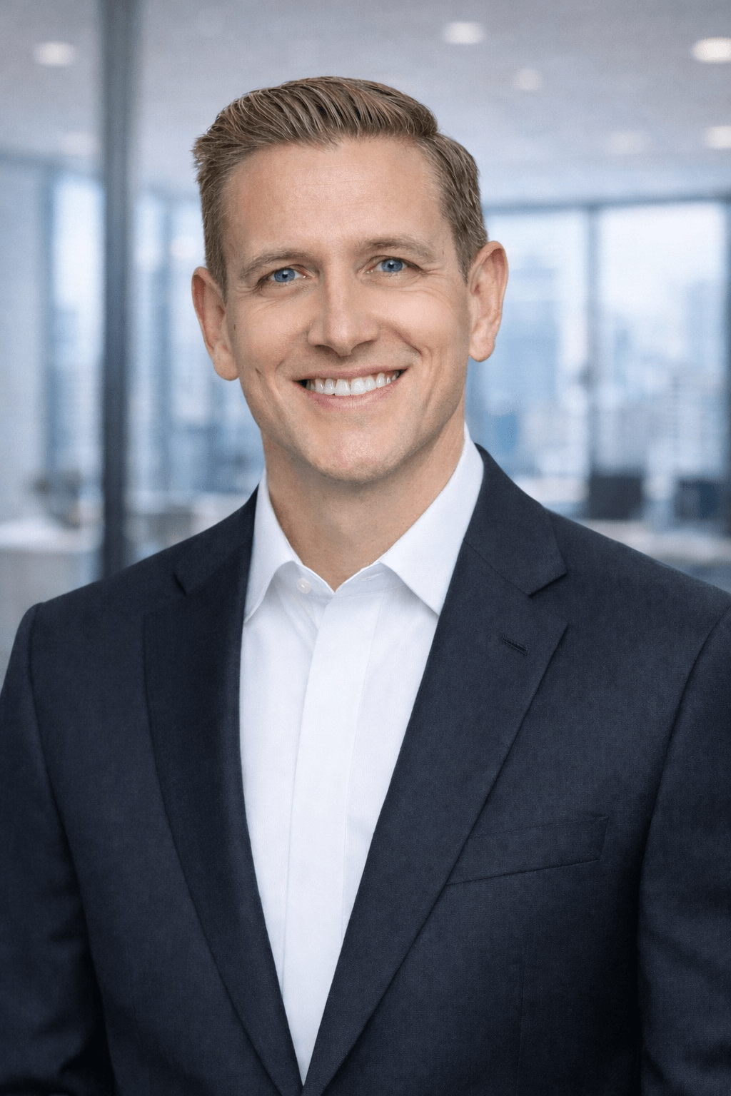 A smiling man in a dark suit and white shirt stands in a modern office setting, conveying professionalism and approachability.